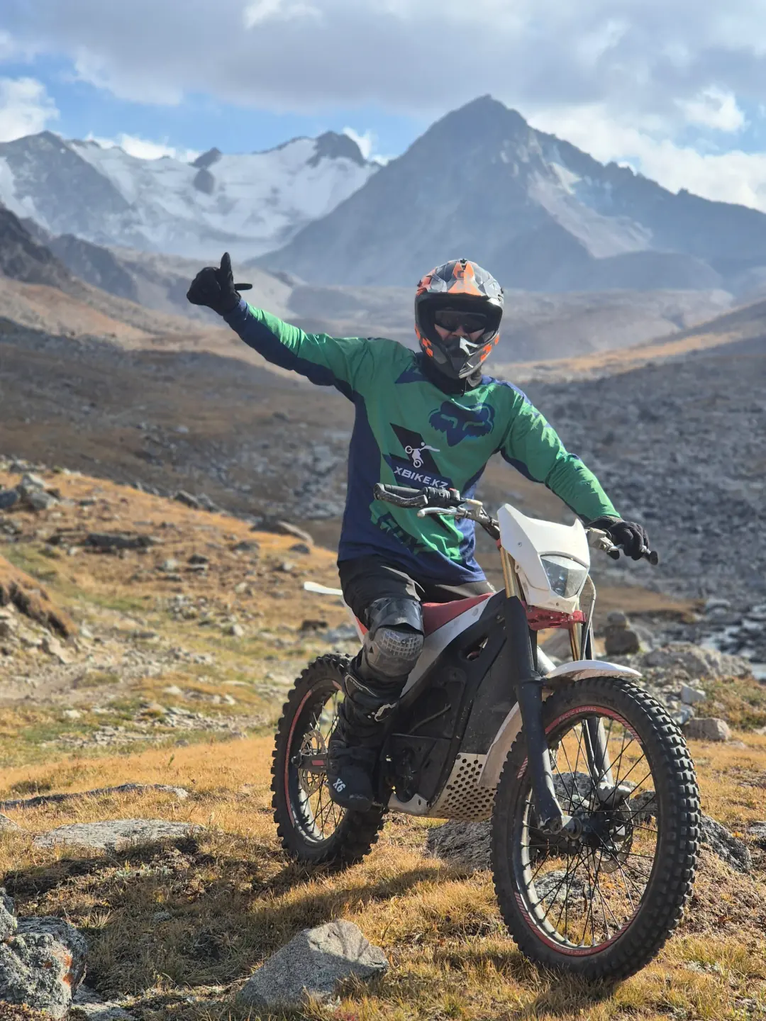 A rider in a green XBIKE.KZ jersey gives a thumbs-up from an electric dirt bike, with snowy peaks behind in Prokhodnoye Gorge.
